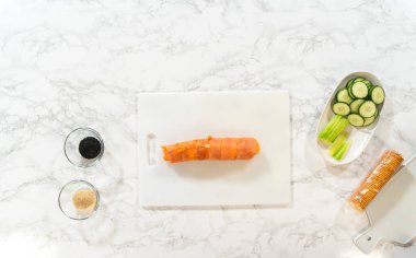 Bıçaklı temiz beyaz bir kesme tahtası, bir blok füme somon, krem peynir, salatalık kurdeleleri, ve siyah-beyaz susam tohumları, hazırlanmaya hazır..