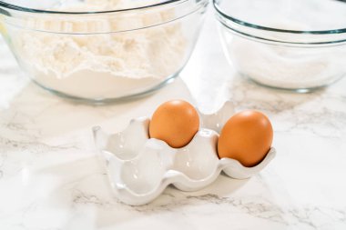 Mermer bir tezgahta seramik yumurta tutacağındaki iki kahverengi yumurtanın açılı görüntüsü. Arka planda, cam karıştırma kâselerinde klasik Sugar Cookie Hamur tarifi için kuru malzemeler var.