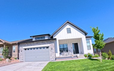 Temiz beyaz bir dış görünüşü, taş aksanları ve parlak mavi gökyüzünün altında gri bir garaj kapısı olan çağdaş bir banliyö evi. Ön bahçede yeşil bir çimen ve küçük ağaçlar, iki sallanan sandalye yer almaktadır.