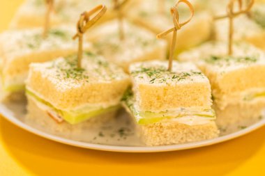 Krem peynir ve salatalık dilimleri ile doldurulmuş, kurutulmuş dereotla kaplı ve bambu kazmalarıyla korunmuş yumuşak beyaz ekmek kareleri. Bu ısırık büyüklüğünde sandviçler beyaz bir tabakta, parlak bir tabakla hazırlandı.