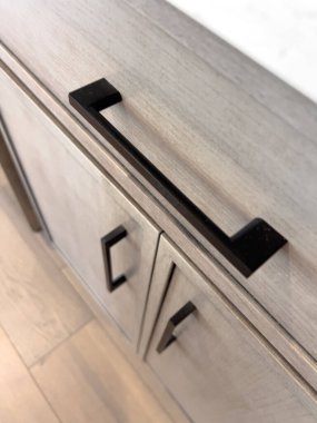 Hafif ahşap bir banyoya monte edilmiş mat siyah çekmece sapı. Yakın plan, yumuşak tonlu fayans zeminine karşı modern tasarımı gösteriyor. Işıklar parlak ve doğal..