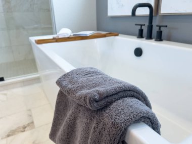 Yan tarafına katlanmış gri bir havlu sarılı beyaz, modern bir banyo küveti. Küvet mat siyah armatürlere sahiptir ve kenarda ahşap bir banyo taşıyıcısı ile cilalanmış mermer zemin üzerine kuruludur..