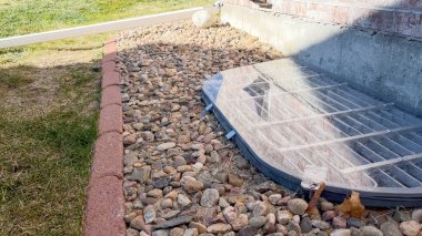 Tam tepeden aşağı bir görünüm, bir evin tuğla temel duvarı boyunca parantez ile korunan şeffaf plastik bir pencere kapağını gösterir.