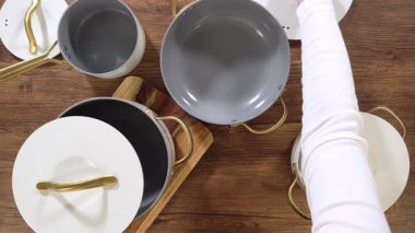 Bir insan eli, altın saplı modern bir seramik tencerenin kapağını kaldırmak için uzanır. Sahne, yumuşak ışıklandırmalı ahşap bir yüzeyde sahneleniyor..