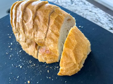 Birkaç dilim dilimlenmiş altın sarısı ekmek, siyah bir yüzeye gevşek bir şekilde dizilmiş. Ekmeğin çıtır çıtır bir kabuğu ve dağınık kırıntıları var. Doğal mutfak ışığı güçlendirir