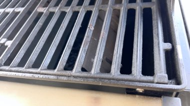 Barbekü ızgaralarının yakın plan çekimi, görünür siyah lekeler ve yağlı birikim. Çekim, iyi aydınlatılmış bir ortamda metal pişirme çubuklarına odaklanıyor..