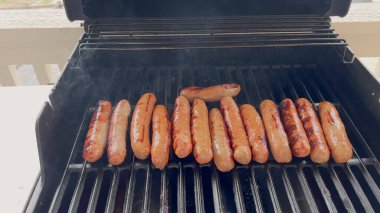 Sıcak bir günde açık havada pişen eşit aralıklı sosislerle dolu bir ızgara. Kapak açık ve sosisler altın kahverengidir. Klasik bir barbekü anını yakalar..