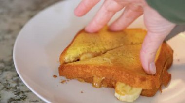 Modern beyaz bir mutfağın şık atmosferinde ızgara fıstık ezmeli ve muzlu sandviç ustalıkla dilimlenir ve el değmemiş beyaz bir tabakta sunulur..