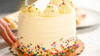 Bir bıçak, üzerinde gül ve gökkuşağı parçacıkları olan bir kutlama pastasını keser. Pasta beyaz mermer bir yüzey üzerinde altın bir tahtanın üzerinde duruyor, neşeli bir anı yakalıyor..