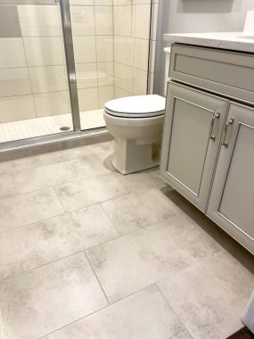 Banyo içi gri dolaplar, beyaz bir tuvalet ve nötr renkli fayans döşemeleri gösteriyor. Duş, modern düzeneği tamamlayan cam bir kapı içeriyor..