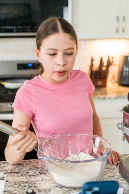 Pembe tişörtlü ve mavi şortlu bir kız mutfak tezgahında kuru malzemeleri karıştırıyor. Mutfak modern ve ışıl ışıl, öğleden sonra pişirme seansı öneriliyor..