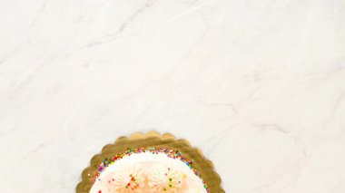 Yuvarlak bir doğum günü pastasını yukarıdan bakıldığında beyaz kremalı ve üzeri şekerle kaplı simetrik şekerlemelerle gösteriliyor. Pasta mermer bir yüzeyde duruyor. Elleri ve aletleri yok..