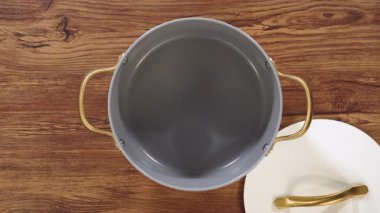 Bir insanın eli, seramik bir tencere kapağının altın sapını yukarıdan tutar. Sahne ışıl ışıl ve ahşap bir yüzey üzerine kurulmuş..