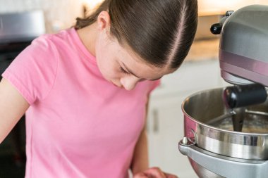 Pembe tişörtlü bir kız yemek hazırlığı sırasında fındıkları karıştırma kabına koyar. Mutfak tezgahı doğal ışık altında malzemeler ve çatal bıçaklarla güzelce düzenlenmiş..