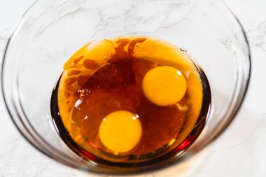 İki çiğ yumurta ve akçaağaç pekmezi içeren cam kasenin üst görüntüsü Amber Zencefilli Kurabiye Hamuru için koyu bir temel karışımı oluşturuyor..