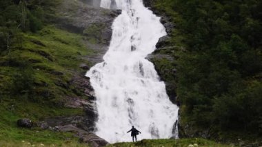 Yağmurluk giymiş bir turist fotoğrafçısı, şelaleli bir uçurumun geniş bir zemininde çelişkili bir şekilde görülebilir..