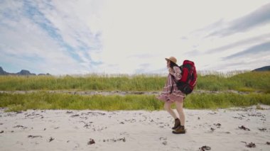 Büyük kırmızı sırt çantalı ve şapkalı bir turist kız dağlarda sahil boyunca yürüyor. Güzel Norveç yaz manzarası