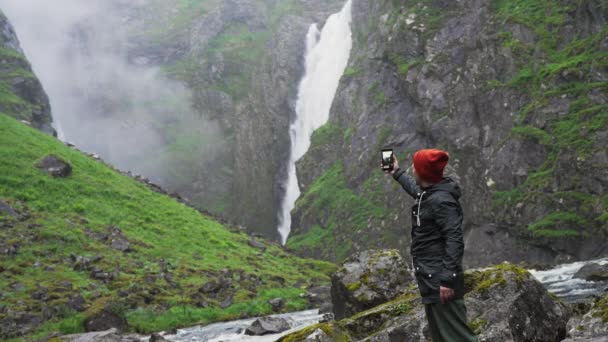 秋のノルウェーの滝の近くに若いカップルのハイキング 岩の上を歩き 景色を眺めながら抱擁する — ストック動画