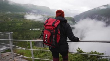Büyük kırmızı sırt çantalı kız gözlem güvertesinde duruyor ve Norveç 'in şelale vadisine bakıyor.
