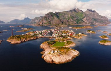 Norveç 'in Lofoten adalarındaki Henningsvaer köyü, futbol sahası ve deniz fenerinin havadan görünüşü bulutlu bir gökyüzünün altındaki kayalık adalar ve dağlarla çevrili.