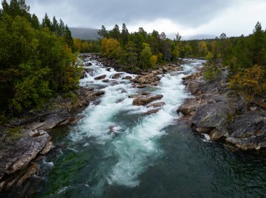 Sükûnet dolu bir nehir, yukarıdaki güzel bulutlu gökyüzünün altında, canlı sonbahar yaprakları arasında zarifçe süzülüyor.