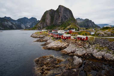 Sakin bir deniz kıyısında canlı kırmızı kulübeler, muhteşem dağlar arka planda güzel bir şekilde siluetlenmiş.