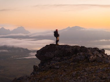 Tek başına bir yürüyüşçü gururla dik bir kayanın tepesinde dikilir ve önlerindeki muhteşem gün batımı manzarasına bakar.