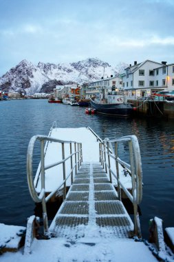 Nefes kesici bir kış manzarası sergiliyor. Etrafı karla kaplı dağlar ve sakin bir limanda tekneler ile çevrili.