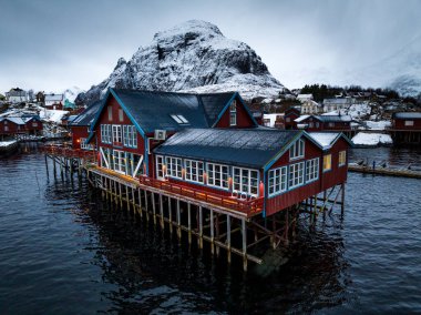Karla kaplı dağlar ve dingin sularla güzel çerçevelenmiş, ayaklıklar üzerinde yükseltilmiş büyüleyici kırmızı bir balıkçı kulübesi keşfedin.