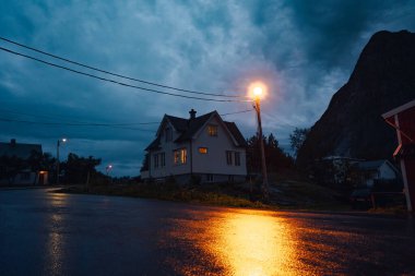 Huzurlu bir gece, rahat bir ev, hafifçe aydınlatılmış, dramatik bulutlar, ve ıslak bir yolda çarpıcı yansımalar.