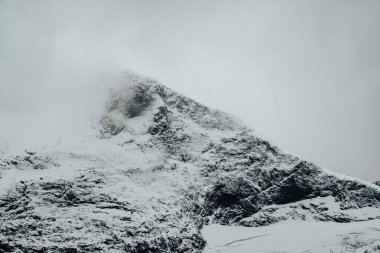 Yumuşak bulutlarla kaplanmış, karla kaplı, sisli bir dağ zirvesini gösteren nefes kesici bir manzara.