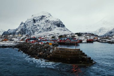 Nefes kesici, karlı bir limanda görkemli, karla kaplı tepelerle çevrili renkli binalar vardır.