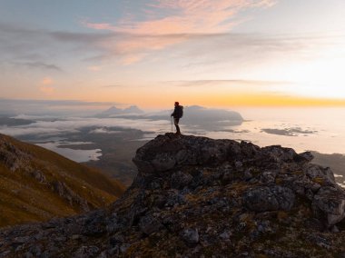 Gün batımında görkemli dağların çarpıcı manzarası, kayalık bir çıkıntıda yalnız bir yürüyüşçüyü barındırıyor, güzelliği kucaklıyor.
