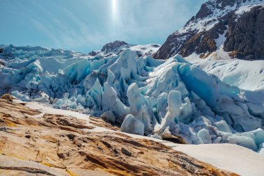 Nefes kesici bir buzulun görüntüsü, parlak mavi gökyüzünün altındaki engebeli kayalar arasındaki çarpıcı buz oluşumlarını gözler önüne seriyor.