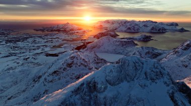 Nefes kesici bir hava manzarası sakin suların üzerinde canlı bir günbatımının yanında görkemli karla kaplı tepeler sergiliyor.