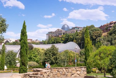 Tbilisi, Georgia - 07 23 2022: Summer view of Rike park in the capital of Georgia Tbilisi Tiflis city with young family walking in front of Rike Concert Hall building surrounded by lush greenery in foreground, and the State Palace of Ceremonies build