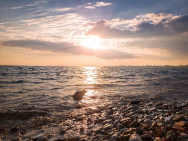 Autumn sunset view across stretch of water with waves beating boulders on the bank under blue sky, sun beaming between pink clouds creating light path on water