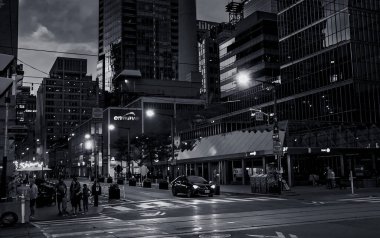 Toronto, Canada - 07 01 2022: Summer night view along Simcoe street with people and a car waiting on traffic loghts in front of glassy walls of scyscrapers reflecting sunset sky in downtown Toronto.
