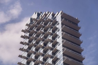 Şehir geometrisi. Yüksek katlı bir binanın tepesinde camsı duvarlar ve balkon kareleri beyaz bulutlarla mavi gökyüzünün önünde bir desen oluşturuyor..
