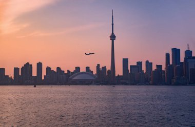 Yaz akşamı manzarası, Ontarios Gölü 'nün iç limanında bir yolcu uçağının Toronto şehir merkezindeki gökdelenlerin siluetlerine karşı kalkışıyla alacakaranlıkta pembe bir gökyüzünün altında.