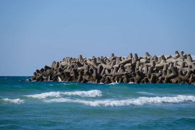 Beton dalga kırıcı tetrapodlar açık mavi gökyüzünün altında kıyı şeridini güçlü deniz dalgalarından koruyor..