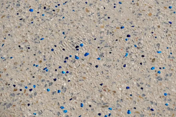 Betona gömülü küçük doğal çakıl taşlarından yapılmış dekoratif bir sahil yolunun yakın çekimi, dağınık mavi cam parçalarıyla. Yüzey, hem işlev hem de estetik olarak tasarlanmış deniz kenarındaki bir yürüyüş yolunun ya da merdivenlerin bir parçasıdır..