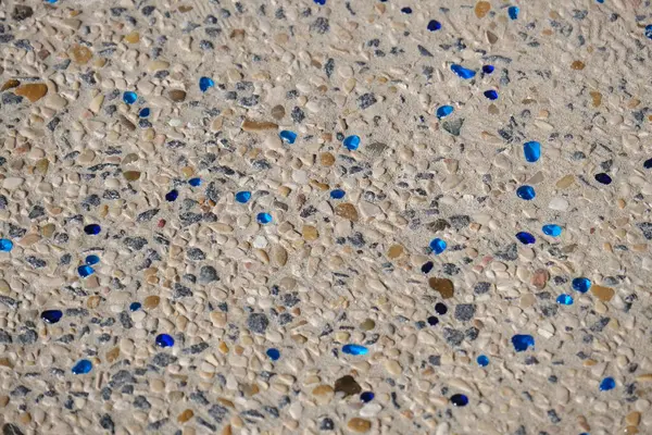 Betona gömülü küçük doğal çakıl taşlarından yapılmış dekoratif bir sahil yolunun yakın çekimi, dağınık mavi cam parçalarıyla. Yüzey, hem işlev hem de estetik olarak tasarlanmış deniz kenarındaki bir yürüyüş yolunun ya da merdivenlerin bir parçasıdır..