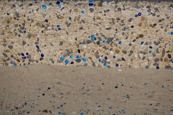 Betona gömülü küçük doğal çakıl taşlarından yapılmış dekoratif bir sahil yolunun yakın çekimi, dağınık mavi cam parçalarıyla. Yüzey, hem işlev hem de estetik olarak tasarlanmış deniz kenarındaki bir yürüyüş yolunun ya da merdivenlerin bir parçasıdır..