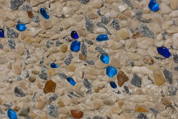 Betona gömülü küçük doğal çakıl taşlarından yapılmış dekoratif bir sahil yolunun yakın çekimi, dağınık mavi cam parçalarıyla. Yüzey, hem işlev hem de estetik olarak tasarlanmış deniz kenarındaki bir yürüyüş yolunun ya da merdivenlerin bir parçasıdır..