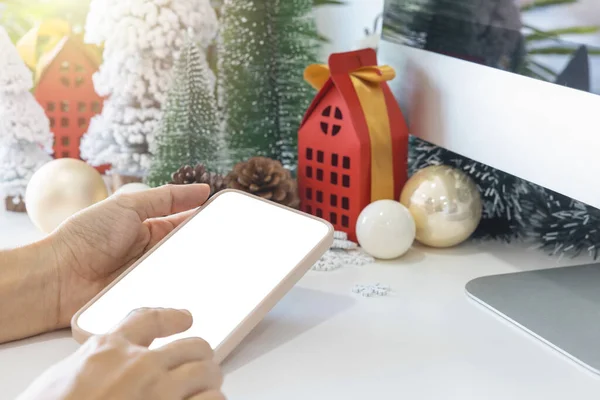 Gentle hands cradle a smartphone with a white screen mock up  by festive scene a beautiful Christmas tree adorned, with Christmas balls, pine cones, and red house gift box tied golden ribbon.