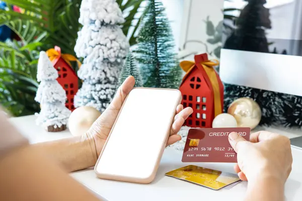 Gentle hands cradle a smartphone with a white screen mock and Hold a credit card to shop online up by the festive scene of a beautiful Christmas tree adorned, with Christmas balls, and pine cones.