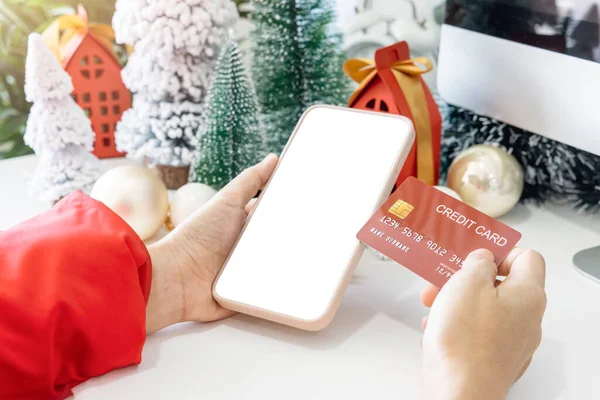 Gentle hands cradle a smartphone with a white screen mock and Hold a credit card to shop online up by the festive scene of a beautiful Christmas tree adorned, with Christmas balls, and pine cones.