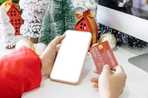 Gentle hands cradle a smartphone with a white screen mock and Hold a credit card to shop online up by the festive scene of a beautiful Christmas tree adorned, with Christmas balls, and pine cones.
