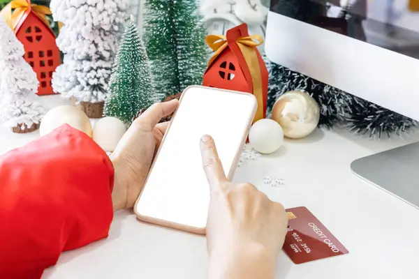 Gentle hands cradle a smartphone with a white screen mock and Fingers on the screen to shop online up by the festive scene of a beautiful Christmas tree adorned, with Christmas balls, and pine cones.